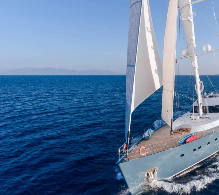 A modern sailboat with white sails glides across deep blue ocean water under a clear sky, with distant land visible on the horizon.