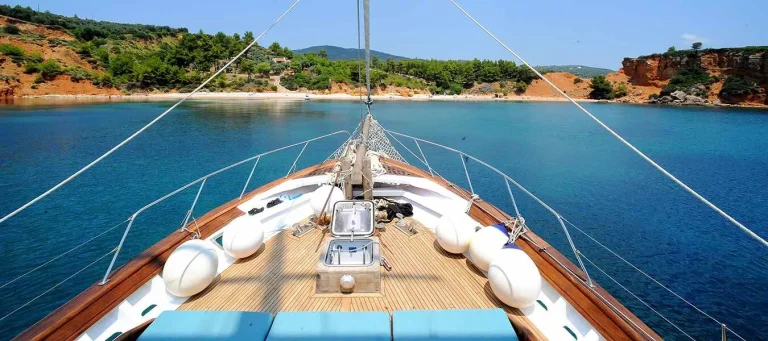 View from the deck of a sailboat approaching a calm, blue bay with tree-lined hills and rocky shores under a clear sky. White railings and lifebuoys line the boat’s wooden deck.