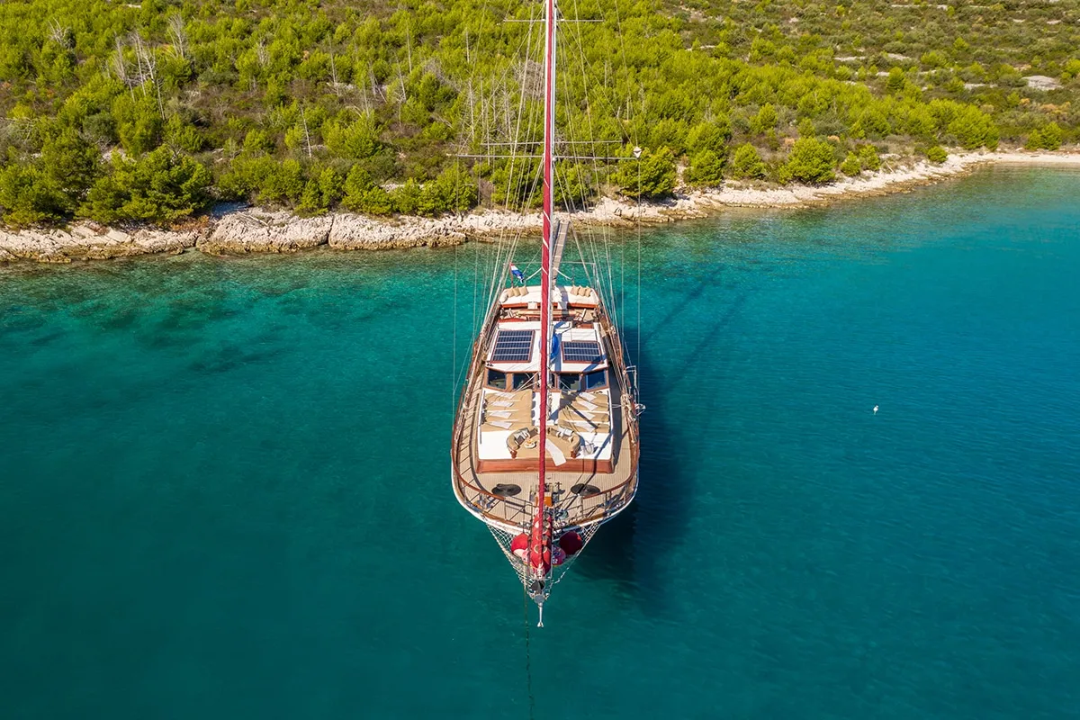 An aerial view of a sailboat anchored in clear turquoise water near a rocky shoreline with dense green trees. The boat has a red mast, visible deck seating, and solar panels.