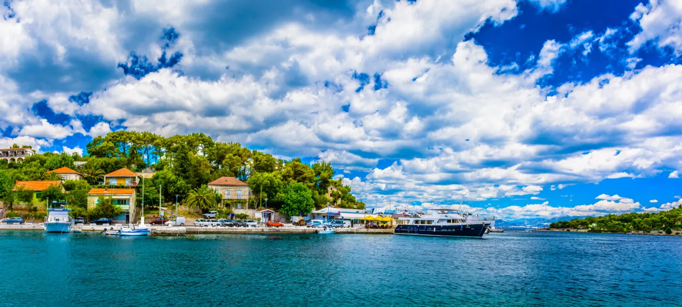 A vibrant coastal village with green trees, colorful houses, and boats docked along the shore under a blue sky filled with dramatic, fluffy white clouds.