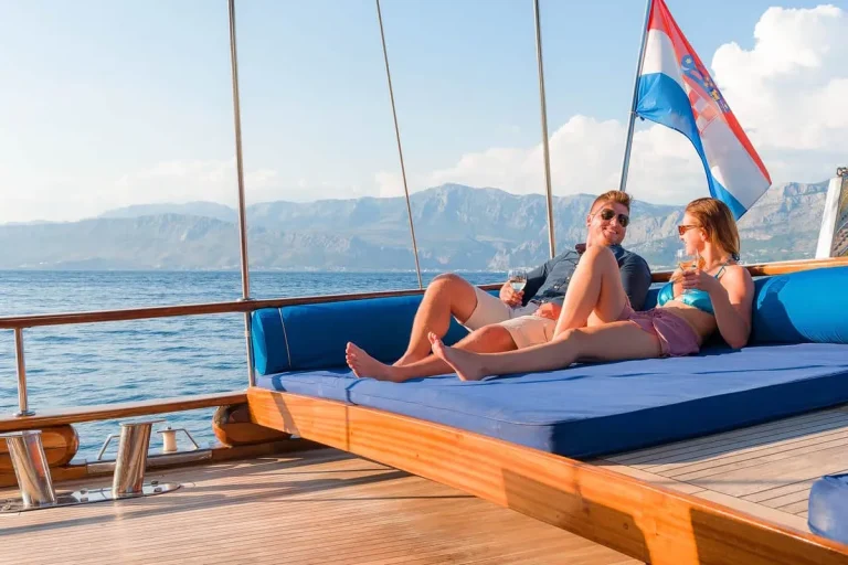 A man and woman relax on a large blue cushioned deck of a sailboat, enjoying drinks. Calm sea and distant mountains are visible, with a Croatian flag waving at the back of the boat. Bright, sunny day.
