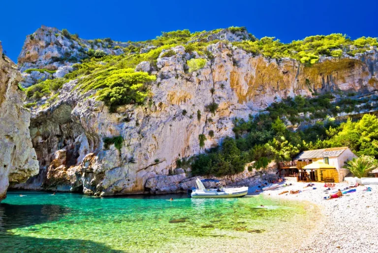 A small beach with clear turquoise water, a white boat, rocky cliffs covered in greenery, and a few people relaxing near a house with a yellow umbrella under a bright blue sky.