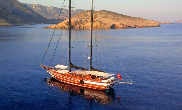 A large wooden sailboat with two masts floats on calm blue sea, near a rocky island with sparse vegetation and distant mountains under a clear sky.
