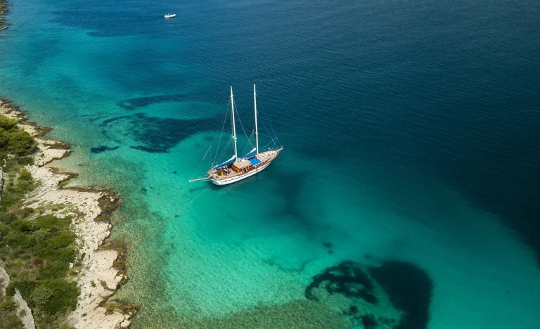 Aerial view of a sailboat anchored in clear turquoise water near a rocky, green shoreline, with deep blue sea extending into the distance and a small white boat further away.