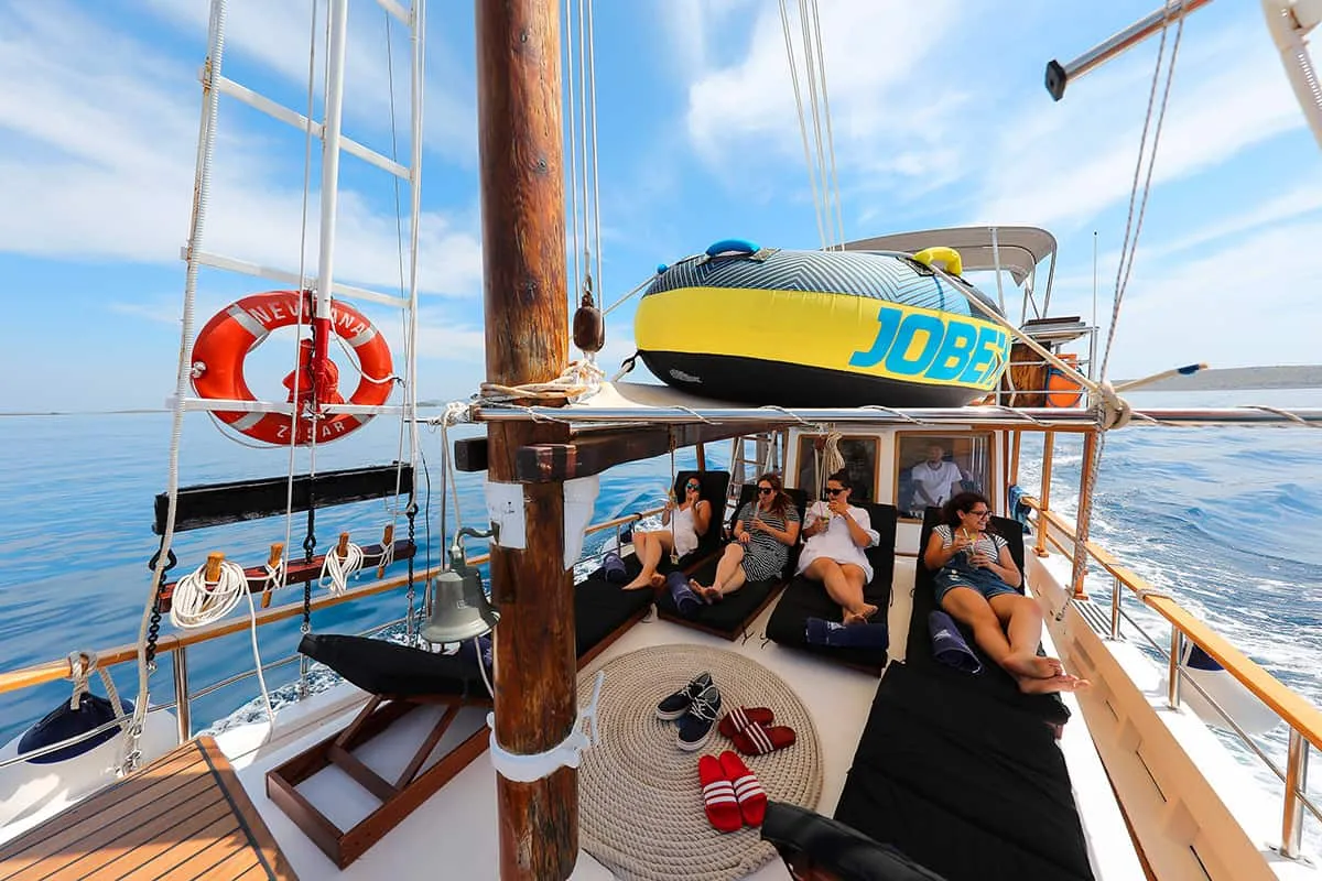A group of people relax on lounge cushions on a sailboat deck under sunny skies, with life rings, ropes, and an inflatable boat visible. Calm blue sea and distant coastline are in the background.