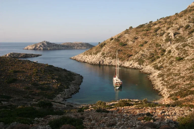A sailboat is anchored in a calm, narrow bay surrounded by rocky hills with sparse vegetation, leading out to the open sea under a clear sky.