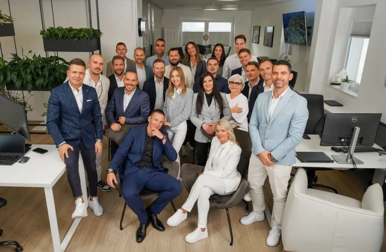 A group of 21 professionally dressed people poses and smiles together in a modern, bright office filled with desks, computers, and plants.