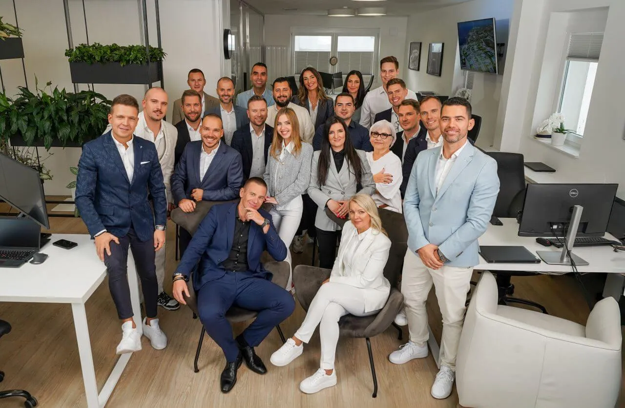 A group of 21 professionally dressed people poses and smiles together in a modern, bright office filled with desks, computers, and plants.