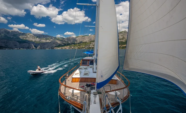 A view from the deck of a sailboat cruising on clear blue water, with white sails raised. A small motorboat passes by on the left, and green mountains are visible in the background under a partly cloudy sky.
