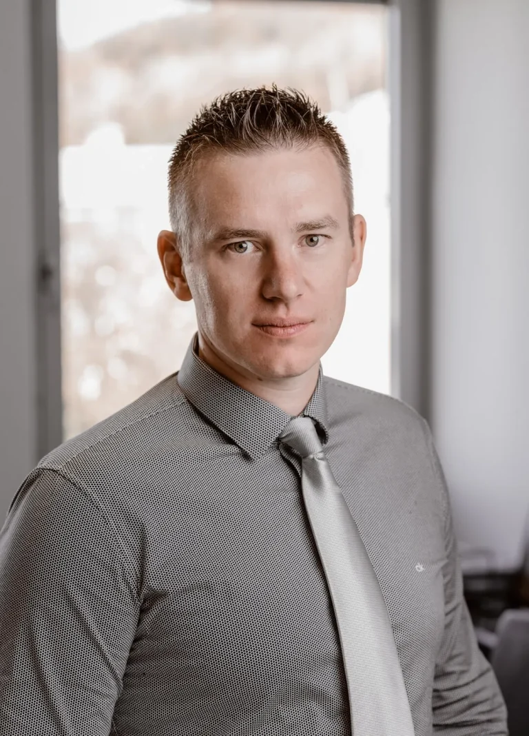 A man with short, light brown hair is wearing a grey dress shirt and a silver tie. He is looking at the camera with a neutral expression, seated indoors with a blurred window in the background.