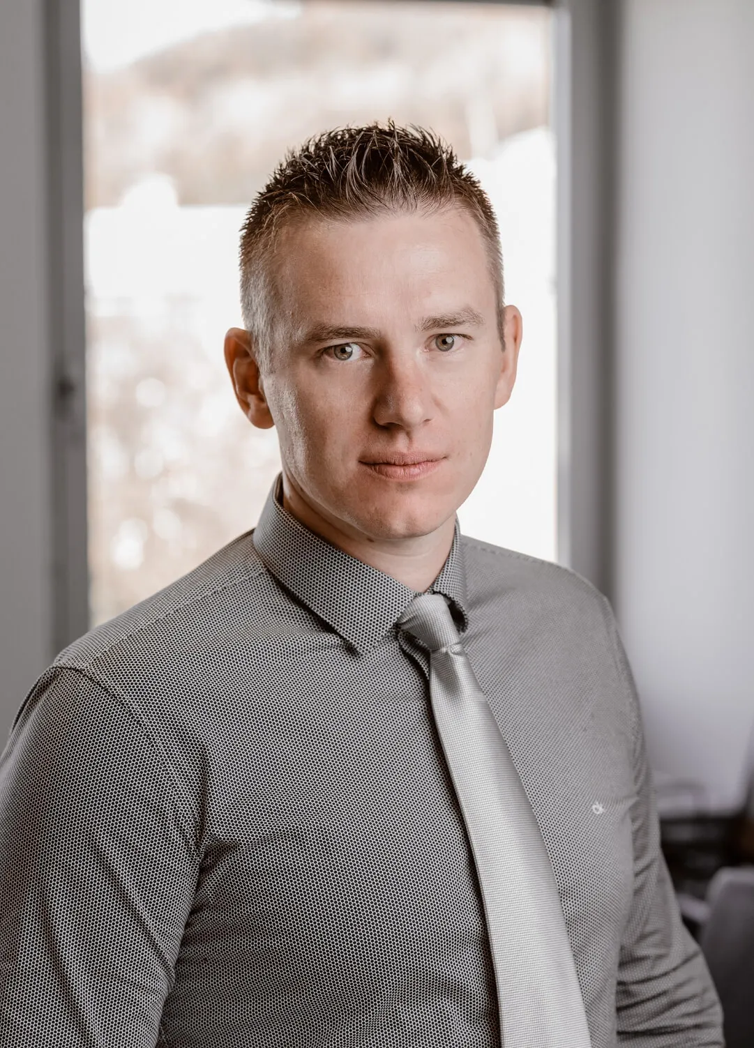 A man with short, light brown hair is wearing a grey dress shirt and a silver tie. He is looking at the camera with a neutral expression, seated indoors with a blurred window in the background.