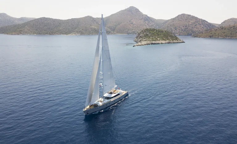 A large sailboat with white sails glides on calm blue water near rocky, green islands and distant hills under a clear sky.