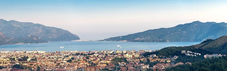 A panoramic view of a coastal city with densely packed buildings in the foreground, surrounded by green hills and mountains with a calm blue bay and boats in the distance under a clear sky.