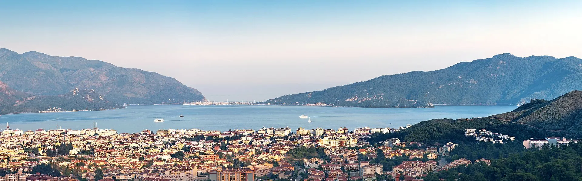 A panoramic view of a coastal city with densely packed buildings in the foreground, surrounded by green hills and mountains with a calm blue bay and boats in the distance under a clear sky.