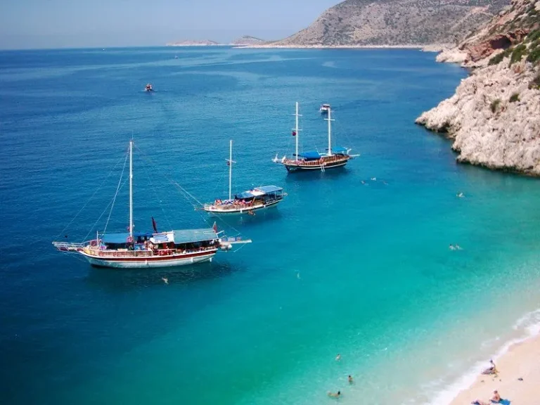 Three sailboats float on clear turquoise water near a sandy beach, with people swimming nearby. Rocky hills and a distant island are visible under a clear blue sky.