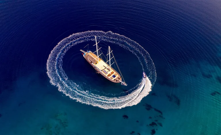 Aerial view of a sailboat floating on clear blue water while a smaller motorboat circles it, creating a white wake pattern around the larger vessel.