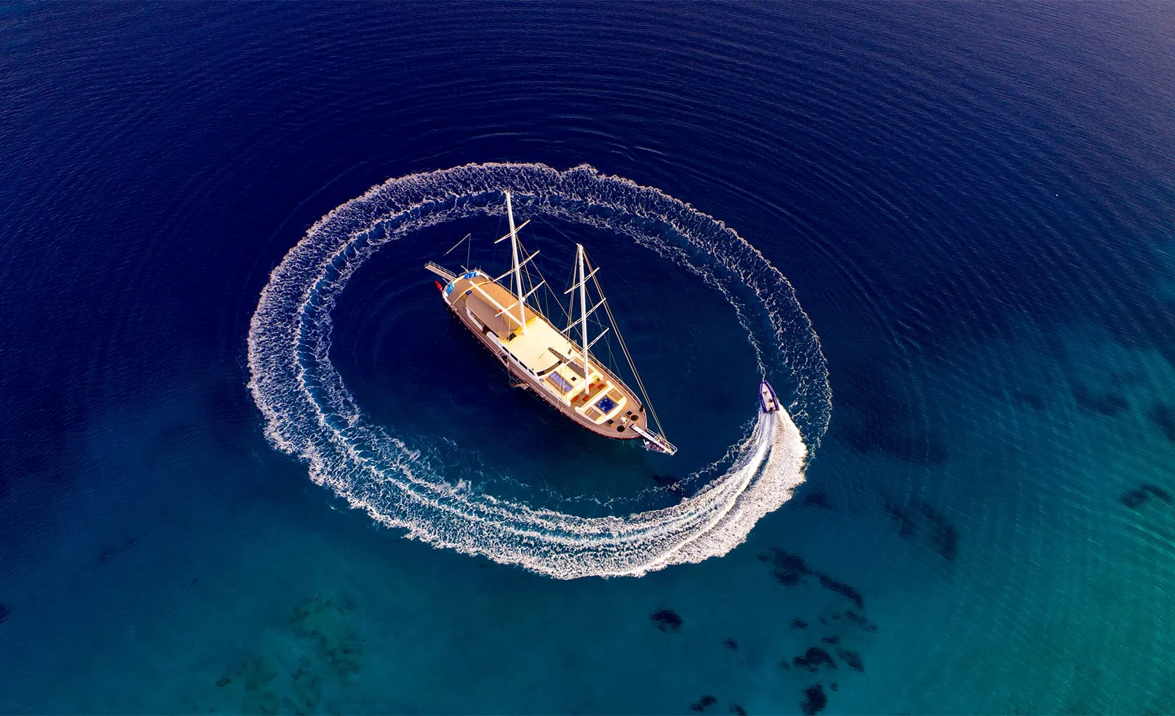 Aerial view of a sailboat floating on clear blue water while a smaller motorboat circles it, creating a white wake pattern around the larger vessel.