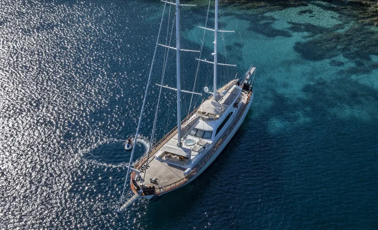 A large white sailing yacht is anchored in clear blue water near a rocky shoreline, with sunlight reflecting off the sea and a small dinghy circling nearby.