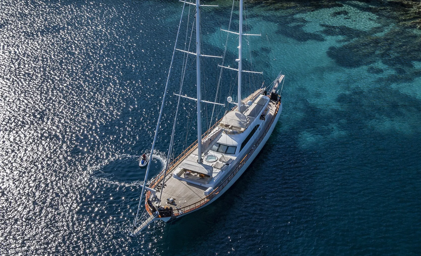 A large white sailing yacht is anchored in clear blue water near a rocky shoreline, with sunlight reflecting off the sea and a small dinghy circling nearby.