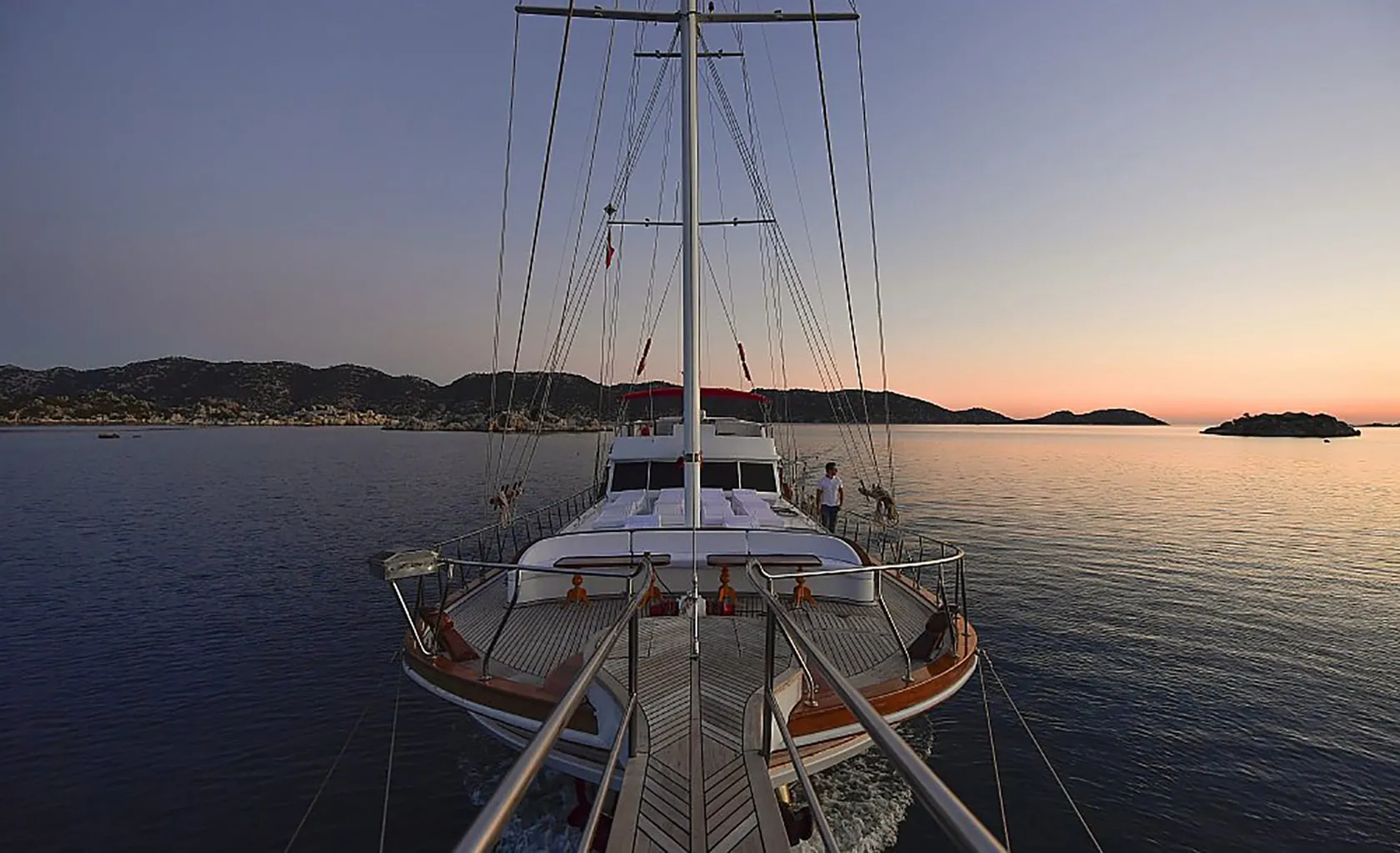 A yacht sails on calm water at sunset, with distant hills on the horizon and a clear sky fading from orange to blue. The photo is taken from the bow, showing the deck and two people on board.