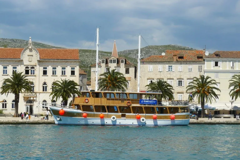 A wooden sailboat with orange buoys is docked by a waterfront lined with palm trees and historic, white stone buildings under a partly cloudy sky.