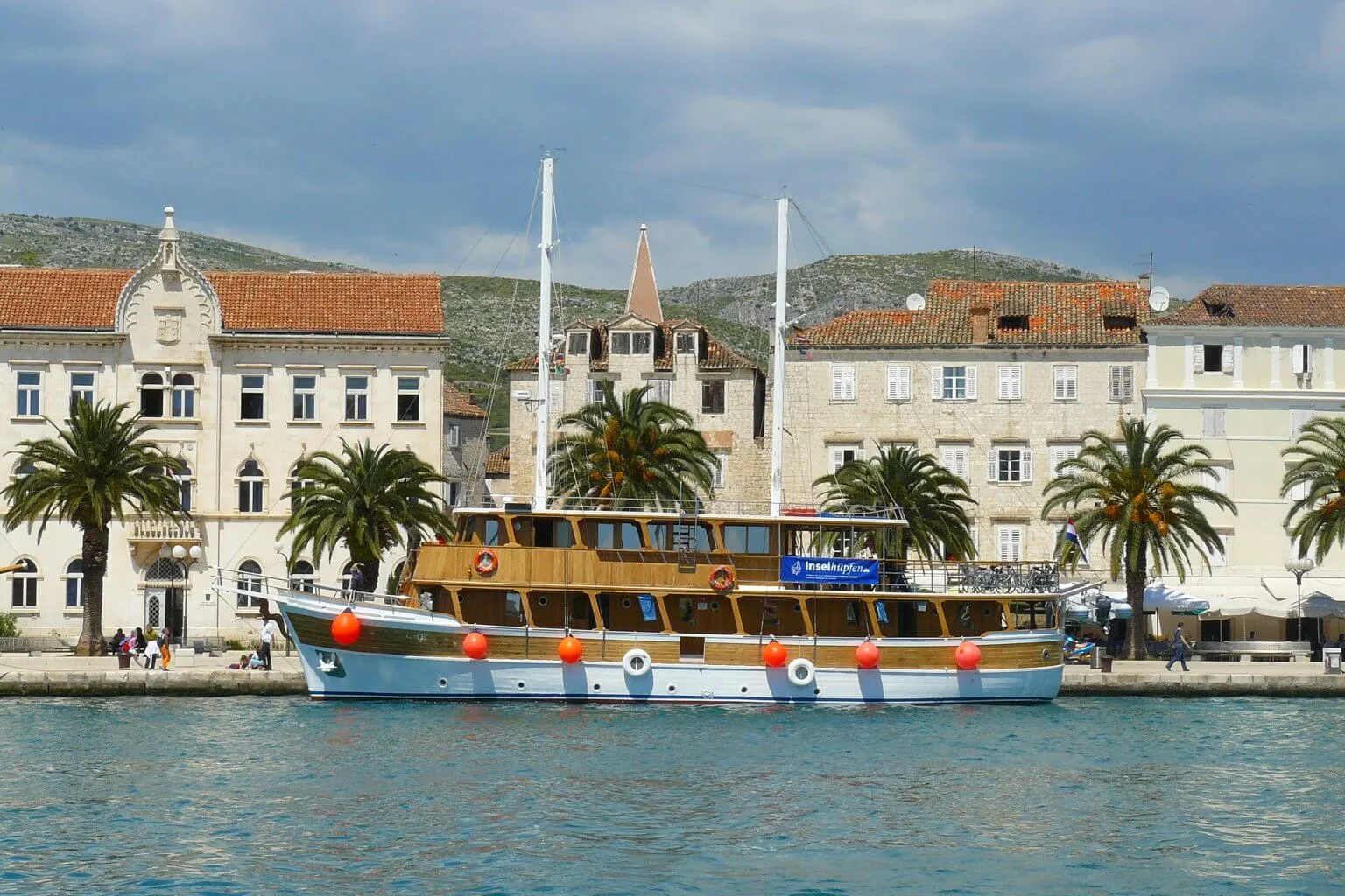 A wooden sailboat with orange buoys is docked by a waterfront lined with palm trees and historic, white stone buildings under a partly cloudy sky.