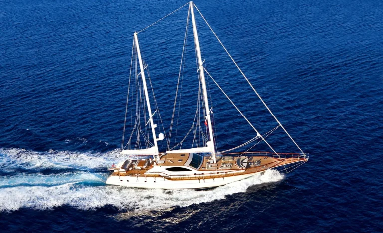 A large white sailing yacht with wooden decking moves swiftly across deep blue ocean water, leaving a foamy wake behind it under a clear sky.
