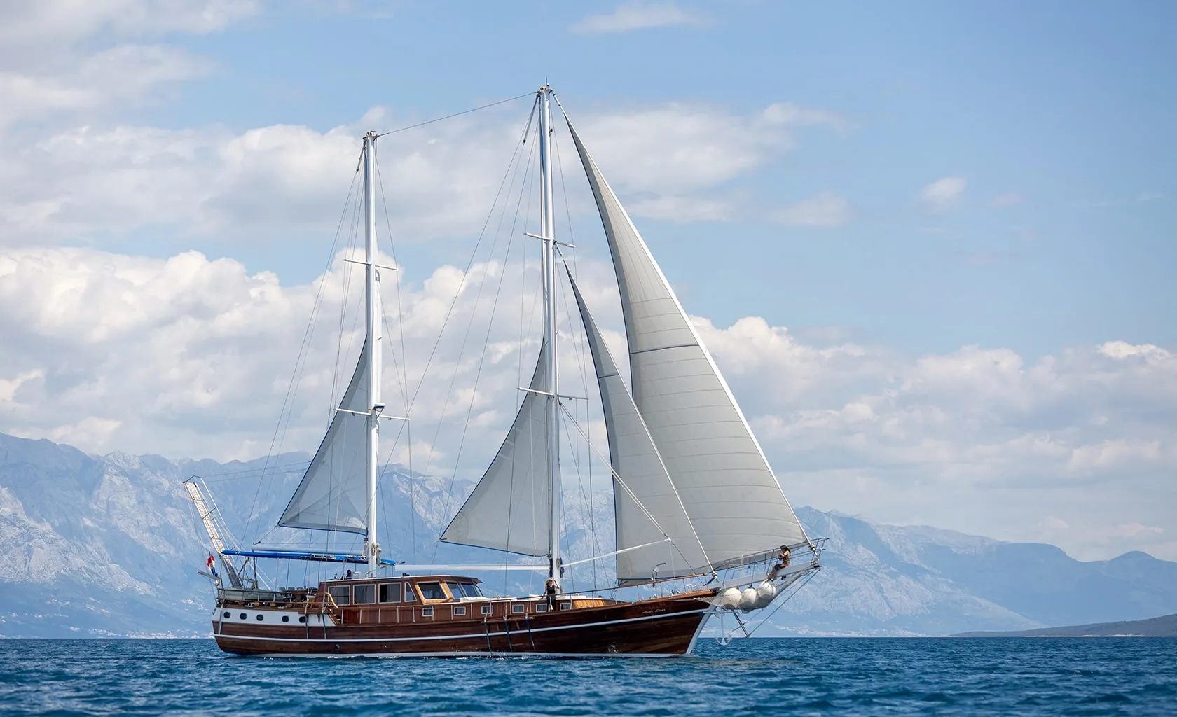 A wooden sailboat with white sails glides on calm blue water, with distant mountains and a partly cloudy sky in the background.