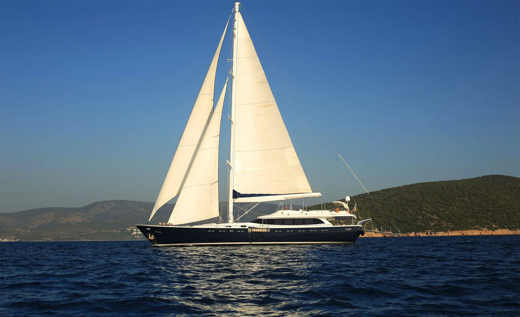 A large sailboat with white sails glides on calm blue water near a coastline with green hills under a clear blue sky.