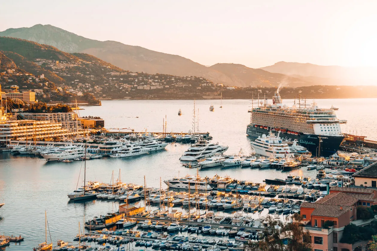 A marina filled with yachts and boats at sunset, with a large cruise ship docked nearby. Hills and buildings surround the harbor, and the sky glows with warm, golden light.