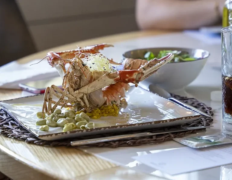 A plated seafood dish featuring a crab presented upright with beans and garnish, served on a square plate. A bowl of salad and a glass of dark beverage are in the background on a wooden table.