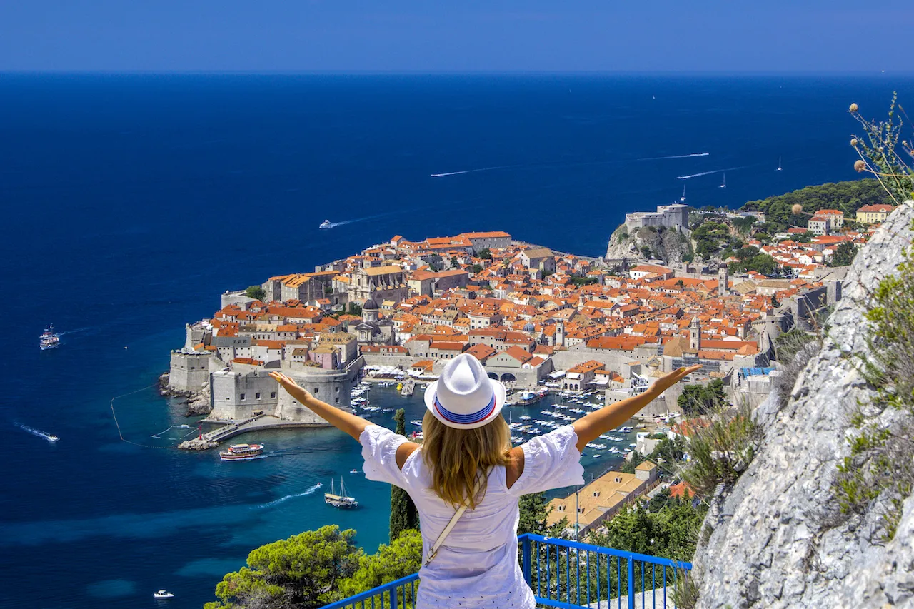 A woman with outstretched arms and a hat stands at a viewpoint, overlooking the historic coastal city of Dubrovnik, Croatia, with its orange rooftops, stone walls, and the deep blue Adriatic Sea in the background.