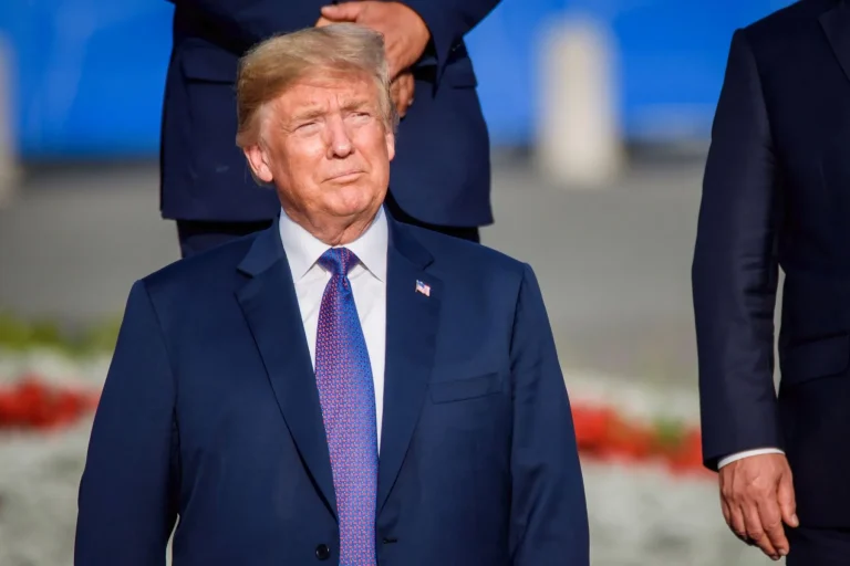 A man in a dark suit and patterned tie stands outdoors, looking slightly upward with a neutral expression. Other people in similar suits are partially visible around him. The background is blurry with blue and red colors.