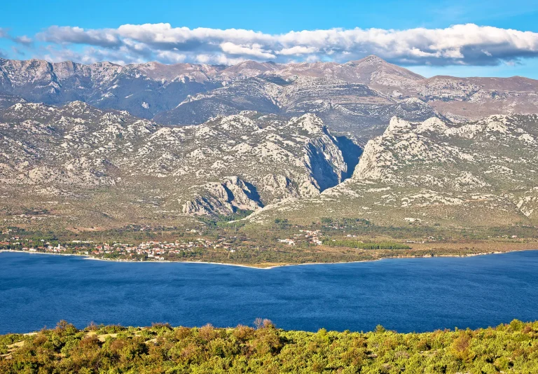A panoramic view of a coastal village beside a deep blue sea, with rocky, rugged mountains and scattered clouds in the background under a bright blue sky.