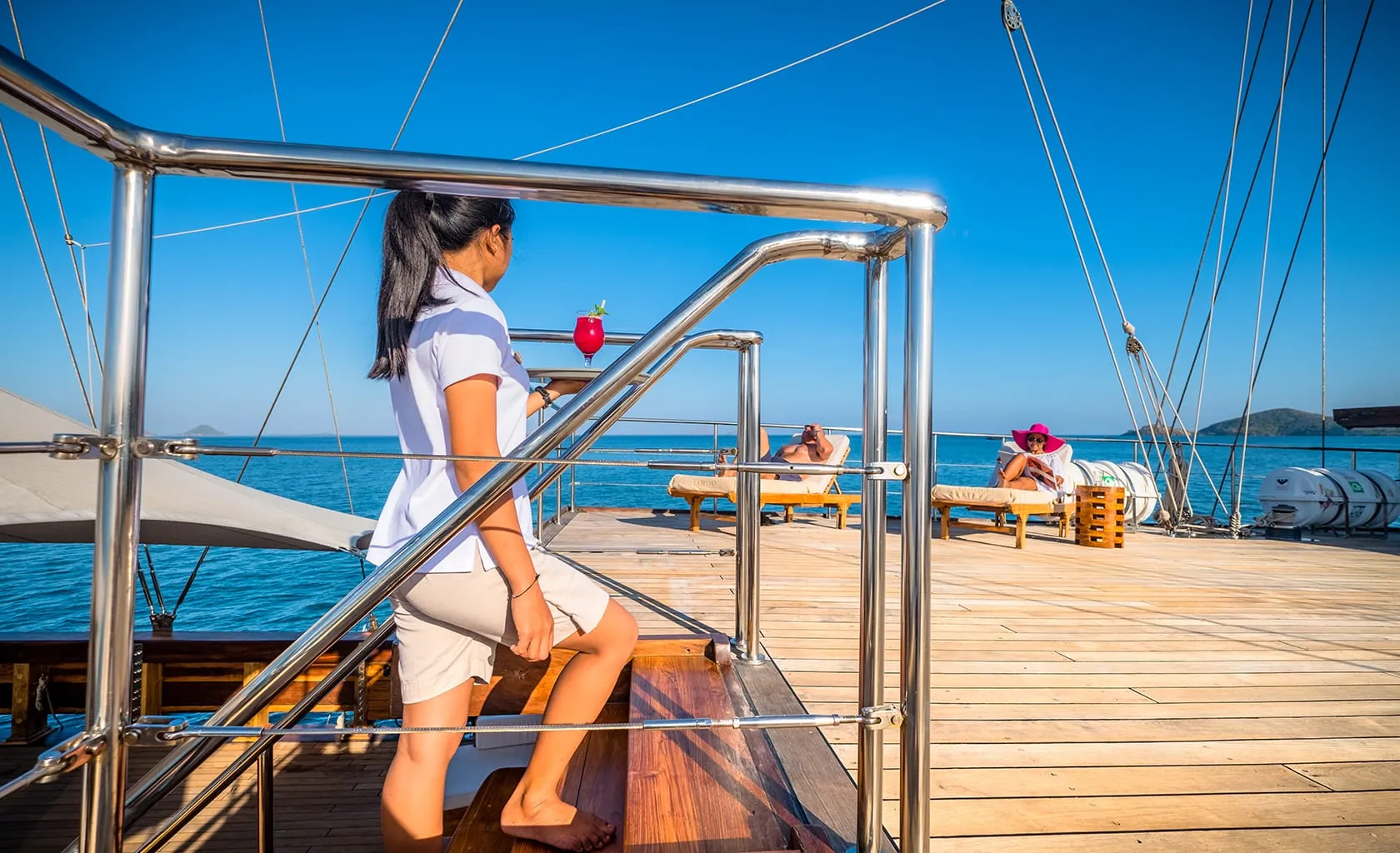 A woman in uniform carries a drink up yacht stairs toward two people relaxing on lounge chairs on a sunny wooden deck, surrounded by blue sea and clear sky.