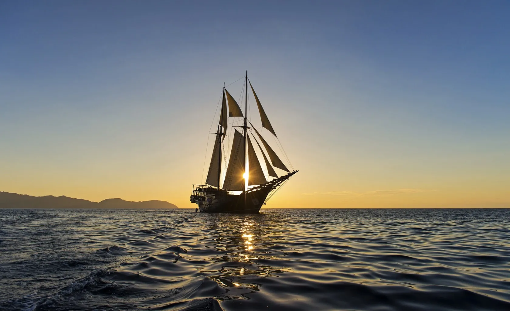 A sailboat with tall sails glides on calm water at sunset, with the sun shining through its sails and distant hills silhouetted on the horizon.