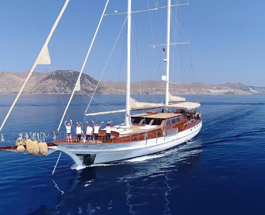A large white sailing yacht with wooden decks moves across calm blue water, with several people standing at the bow waving. Rocky hills and a coastal town are visible in the background under a clear sky.