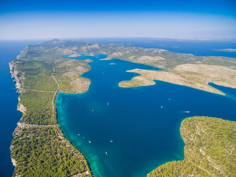 Aerial view of a coastline with clear blue water, several small islands, peninsulas covered with green vegetation, and multiple sailboats scattered in the water under a clear, blue sky.