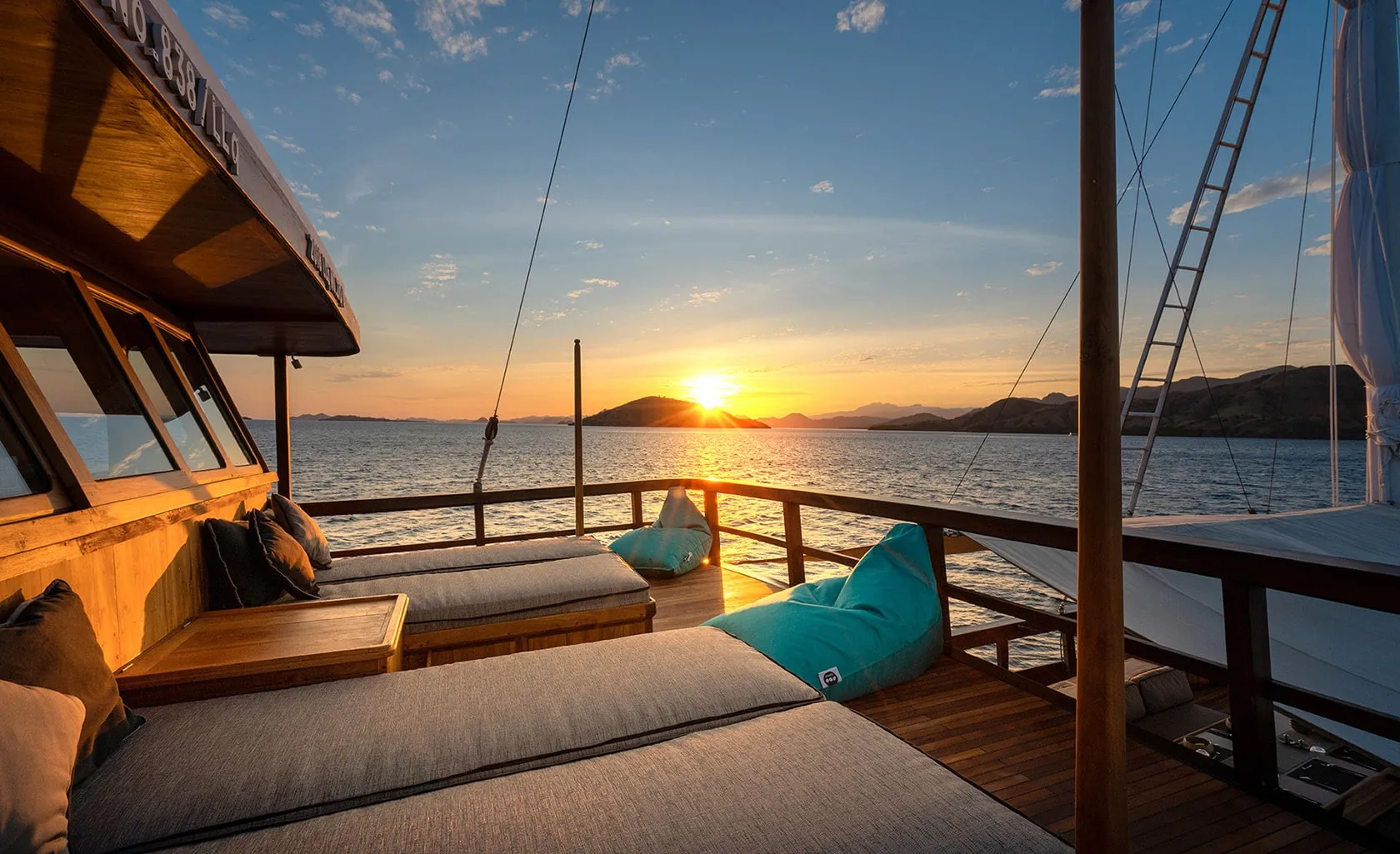 A serene sunset view from the deck of a boat, featuring cushioned lounge chairs and bean bags, overlooking calm water and distant hills under a colorful sky.