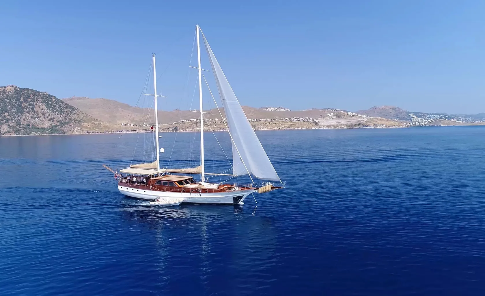 A white sailboat with raised sails glides on calm blue water near a coastline with hills and scattered buildings under a clear, sunny sky.