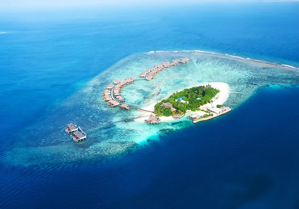 Aerial view of a small tropical island surrounded by turquoise water, featuring overwater bungalows, a dock, and lush greenery at the center, all set in a vast blue ocean.