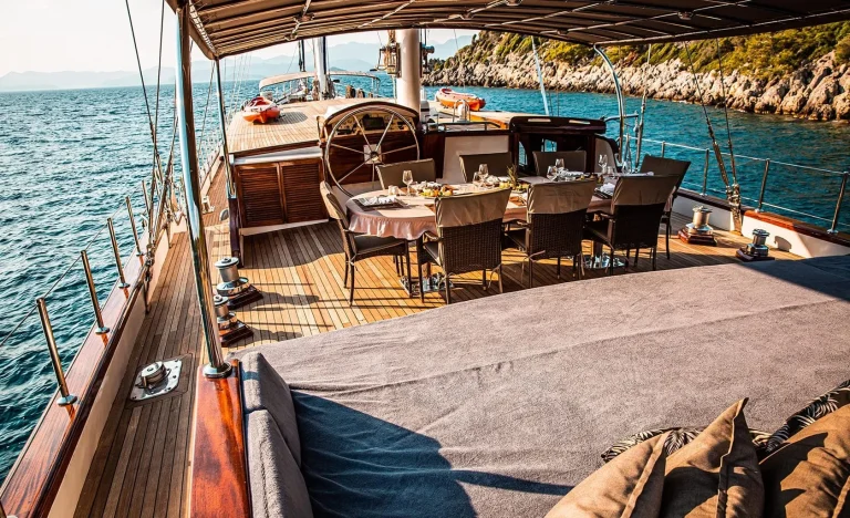 A luxury yacht deck features a dining table set for six, cushioned seating, and a shaded area. The yacht sails near rocky coastline with calm blue water and distant mountains under a clear sky.