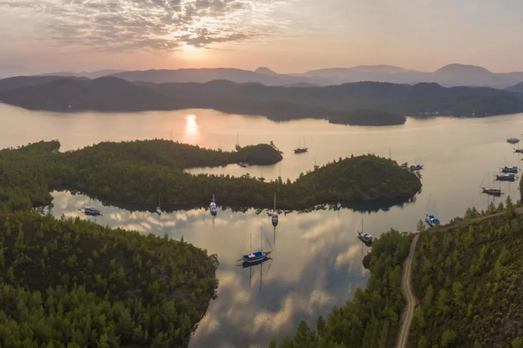 A tranquil bay at sunset with calm water, small forested islands, and several boats anchored. The sky is partly cloudy, and the sun is low on the horizon, casting warm light over the serene landscape.