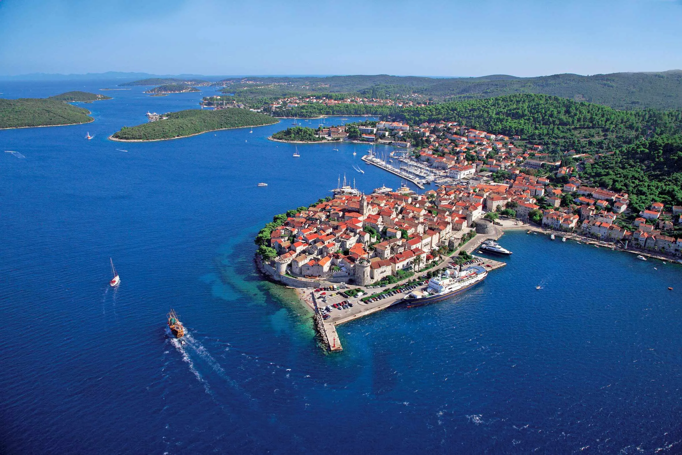 Aerial view of a coastal town with red-roofed buildings, surrounded by blue sea and forested hills. Boats and yachts are docked along the shoreline and sailing in the water.