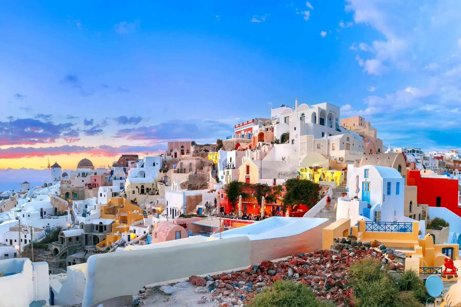 Colorful white and pastel houses with blue domes on a hillside in Santorini, Greece, under a vibrant sunset sky with scattered clouds and the sea visible in the background.