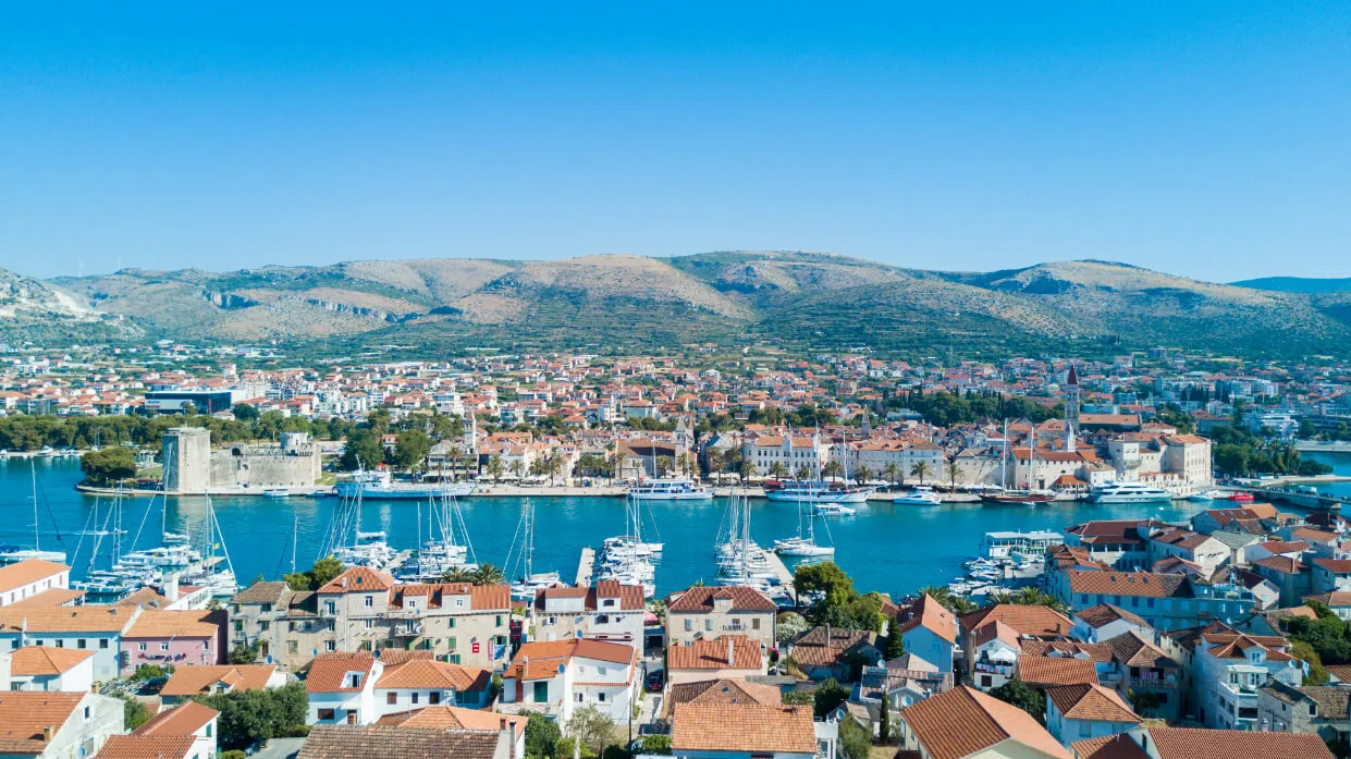 Aerial view of a coastal town with red-roofed buildings, a marina filled with boats, and rolling hills in the background under a clear blue sky.