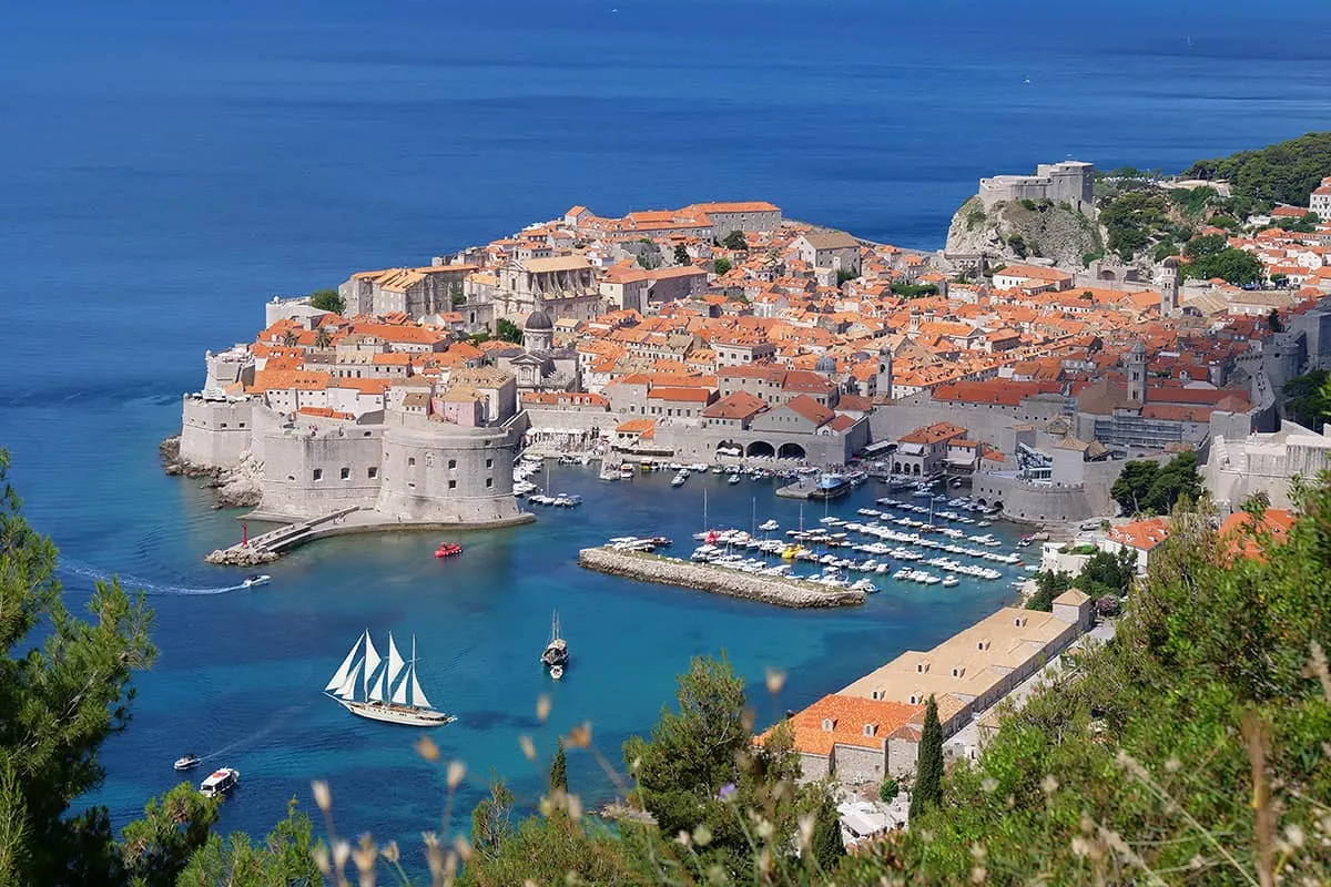 A scenic view of Dubrovnik’s old town, with historic stone buildings and orange rooftops surrounded by city walls, a turquoise harbor with boats, and the blue Adriatic Sea in the background.
