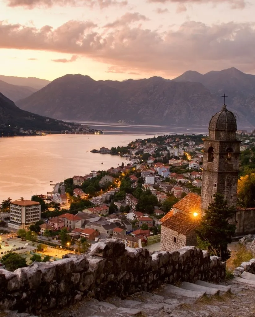 View of a coastal town at sunset with red-roofed buildings, a stone church tower in the foreground, calm water, and mountains in the background under a partly cloudy sky.
