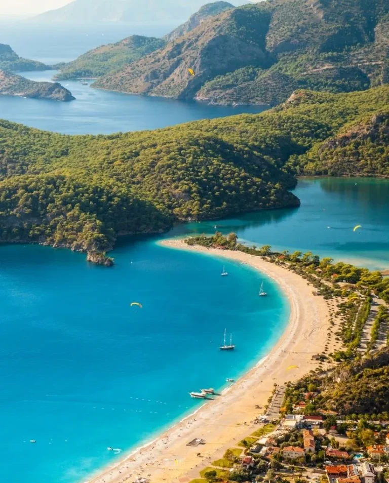 Aerial view of a curved sandy beach with turquoise waters, surrounded by lush green hills and mountains, with boats and parasailers in the water and small buildings lining the shore.