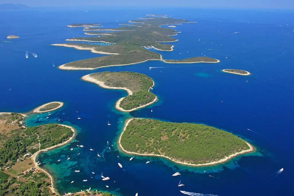Aerial view of a chain of green, tree-covered islands surrounded by bright blue sea, with numerous sailboats and yachts scattered along the coastlines and inlets.